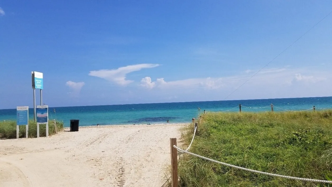 A sandy path opening onto a calm sea-glass green ocean under a blue sky
